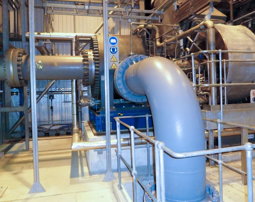 An industrial interior view showing a large horizontal machine, possibly a gearbox or turbine, connected to a substantial, gray bent pipe, surrounded by safety railings and structural piping.