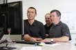 Drei Personen in Arbeitskleidung stehen gemeinsam vor einem Computerbildschirm und betrachten konzentriert die Anzeige; sie wirken, als würden sie technische Daten besprechen oder an einer gemeinsamen Aufgabe arbeiten.