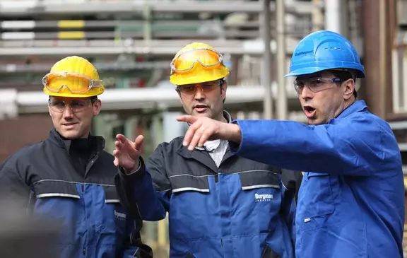 Three industrial workers wearing safety helmets discussing a task on a plant site while pointing toward equipment.