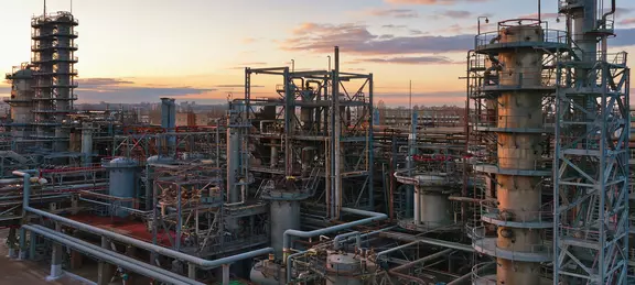 Eine erhöhte Ansicht einer großen, komplexen Industrieanlage für chemische Produkte oder einer Ölraffinerie mit zahlreichen Metallrohren, Lagertanks und Destillationskolonnen vor dem Hintergrund eines Sonnenuntergangs.