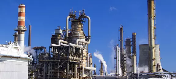 Industrial oil refinery with multiple distillation towers, pipelines, and chimneys emitting steam or smoke under a clear blue sky.