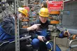 Two industrial technicians wearing protective helmets inspecting and measuring equipment in a complex pipe system, with one pointing toward a valve.