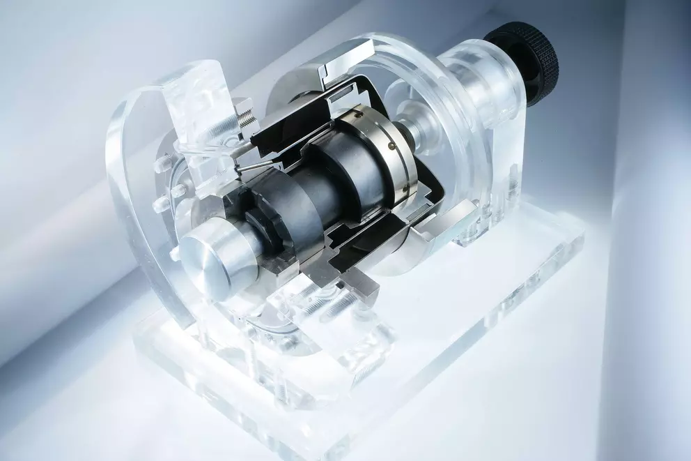 A studio photograph of a cutaway demonstrator model, showing the internal components of a mechanical seal or a small fluid drive, made of clear acrylic, metal, and black plastic parts, mounted on a transparent stand.