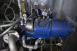 Zwei Ingenieure mit Schutzhelmen inspizieren eine große blaue Industriemaschine, die von Rohrleitungen und Messinstrumenten umgeben ist.