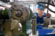 Technician wearing a safety helmet and goggles working on a complex industrial system with pipes, sealing equipment, and measurement instruments.