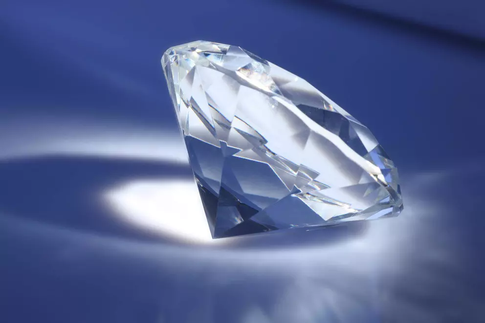 Ein großer Diamant im Brillantschliff auf einer blauen Oberfläche, beleuchtet von einem Scheinwerfer, der seine Facetten hervorhebt und einen hellen Schatten wirft.