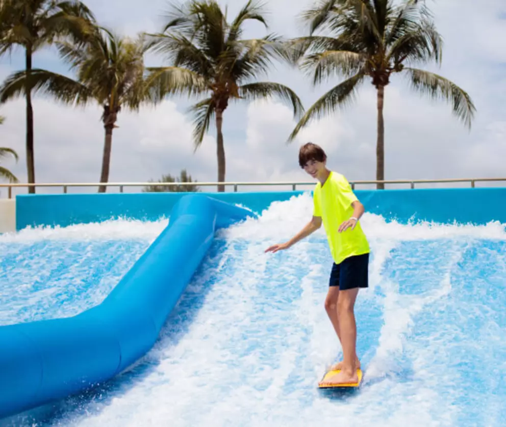 Teenager surfing on an artificial wave at a tropical water park with palm trees in the background.