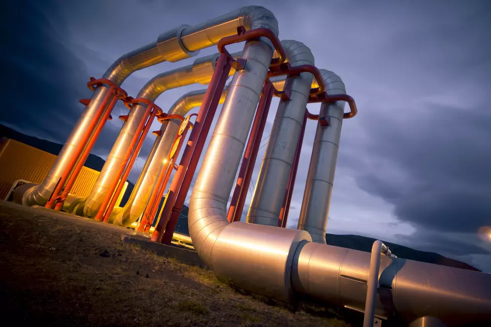 Large, silver industrial pipes with red supports curve upwards against a dramatic, dark blue evening sky.