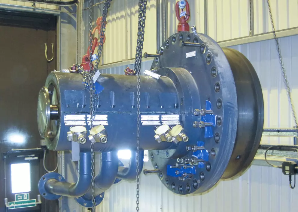 A large, dark industrial machine component, possibly a heavy-duty pump or turbine section with multiple bolted connections and attached piping, is suspended in a factory environment by thick metal chains.
