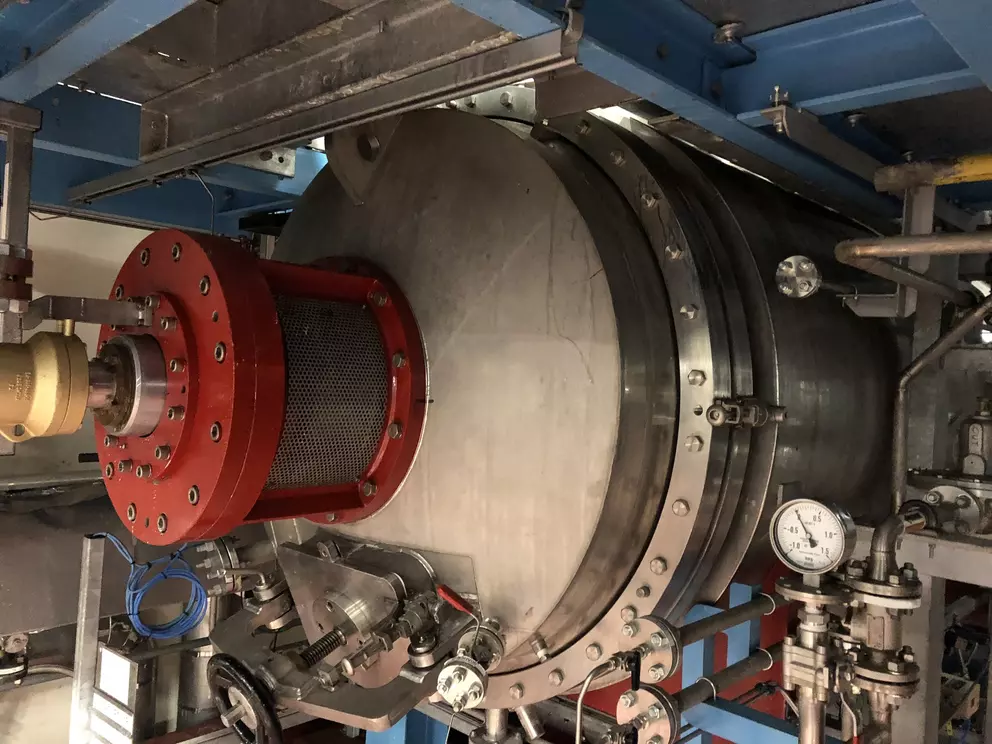Industrieanlage mit einem großen, zylindrischen Behälter aus Edelstahl mit einem auffälligen roten Flanschanschluss und einem Maschenfilter, umgeben von Rohrleitungen und Messgeräten.