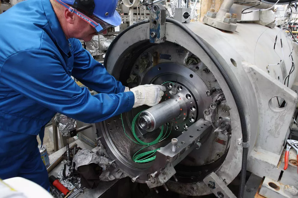 A technician wearing a blue uniform, hard hat, and safety glasses is installing a large component or shaft into a complex piece of industrial machinery, likely a turbine or compressor.
