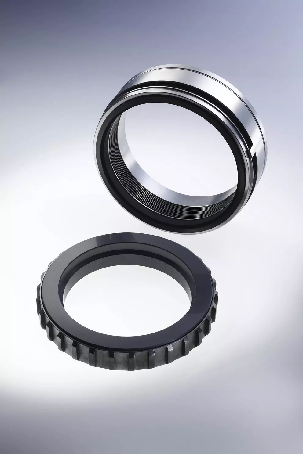 A studio shot of two circular mechanical seal components: a highly polished, silver metal sleeve floating above a darker, toothed seal face ring.