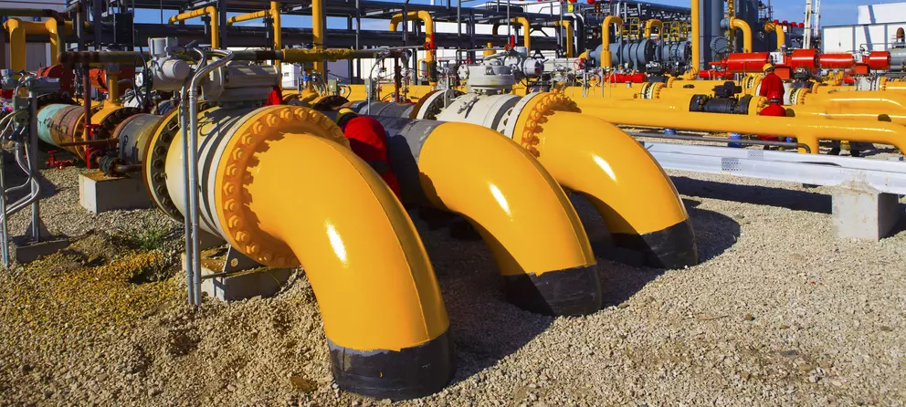 A close-up view of three large, bright yellow industrial pipelines and elbows connected with large flanges, running over a gravel ground at a processing facility, with a network of other colorful pipes and valves in the background.