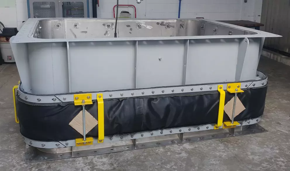 Large rectangular industrial metal casing or duct section with a black flexible expansion joint wrapped around the base.