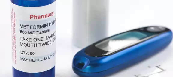 Close-up of a prescription bottle labeled Metformin next to a blood glucose meter and test strips on a white surface.