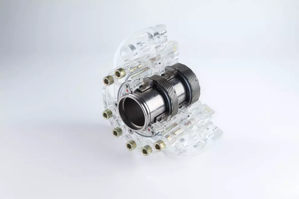A mechanical component, featuring a central metallic cylinder with a corrugated section, encased by a segmented, transparent acrylic housing held together by several small bolts, resting on a white surface.
